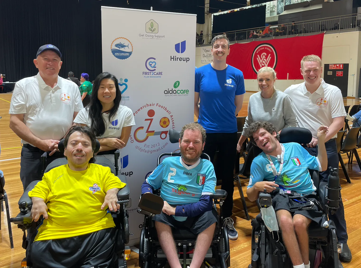 Tristram and his team mates are smiling. Behind them are sponsors of the Queensland powerchair football association from different organisations including Hireup. Between them is a banner with the logos of the different sponsors.