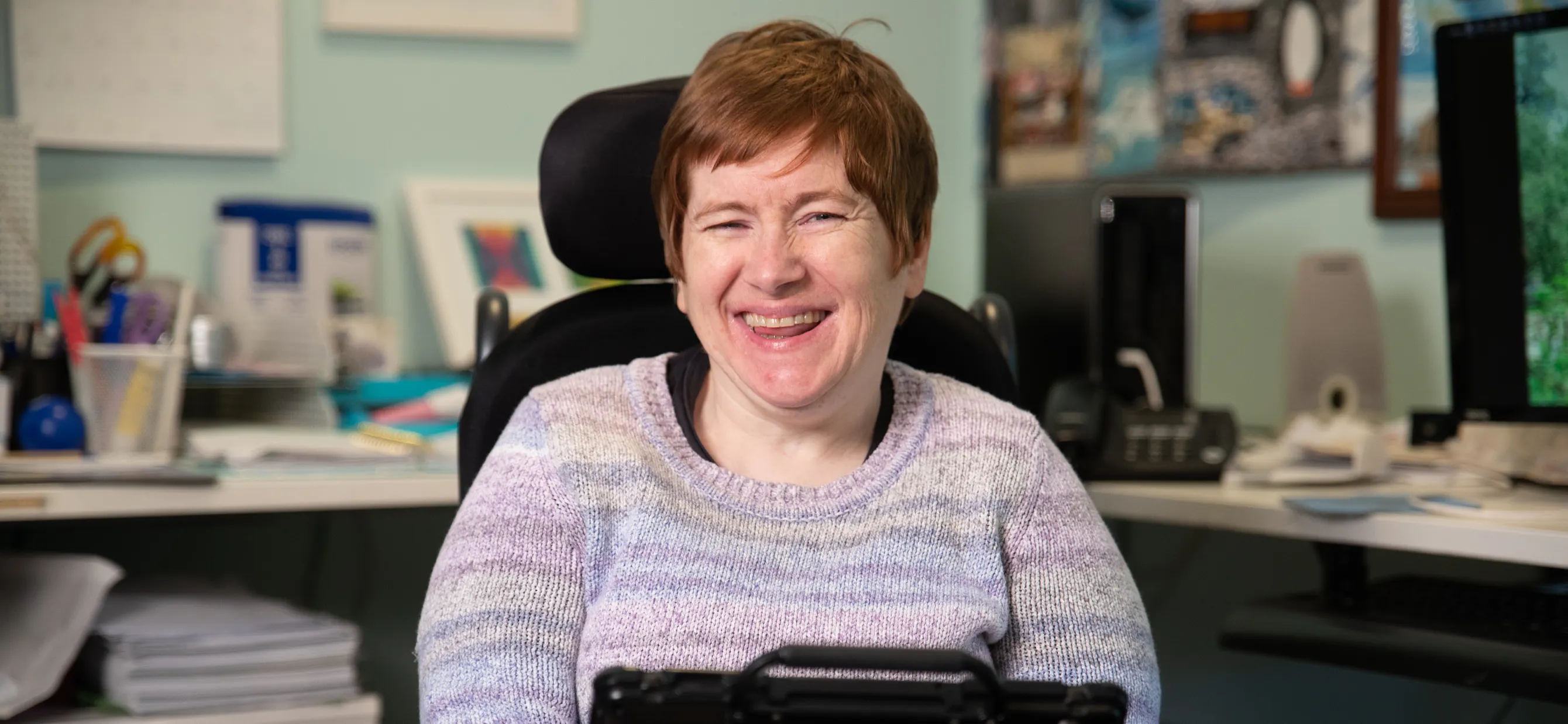 Fiona has red hair and is wearing a purple jumper. Behind her is a home office setting with a long desk, computer desktop and stationary. Fiona is a wheelchair user and has an Alternative and augmentative communication device in front of her. Fiona is smiling. 