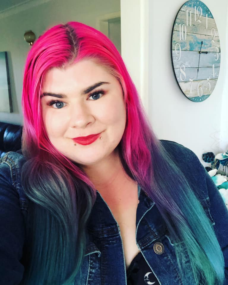 Zoe has long bright pink and aqua hair. She is wearing red lipstick and a blue denim jacket. There is a clock in the background. 