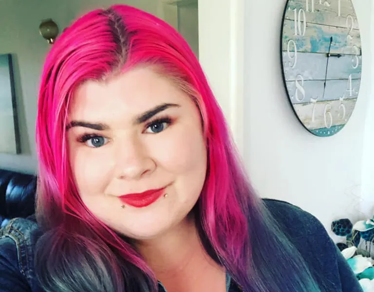 Zoe has long bright pink and aqua hair. She is wearing red lipstick and a blue denim jacket. There is a clock in the background.