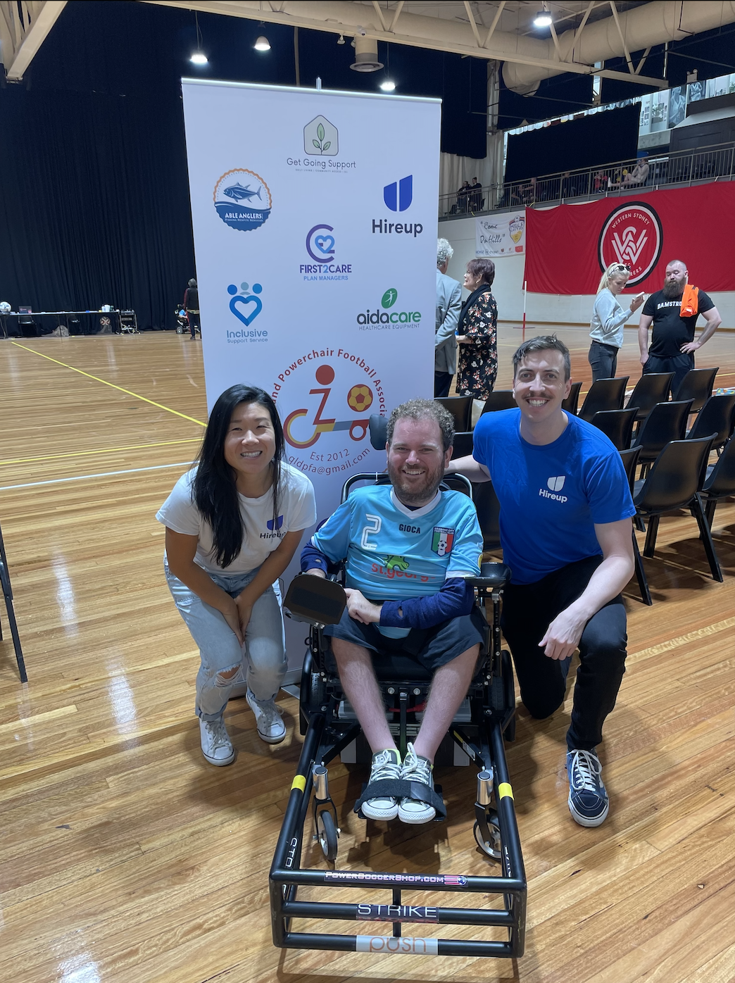 Tristram is between Steph and David from the Hireup support office. Tristram is wearing a light blue and white football jersey. Steph is wearing a white Hireup t-shirt and David is wearing a Blue Hireup t-shirt. There is a banner behind them displaying the sponsors of the Queensland powerchair football association. Tristram uses a wheelchair. Everyone is smiling.