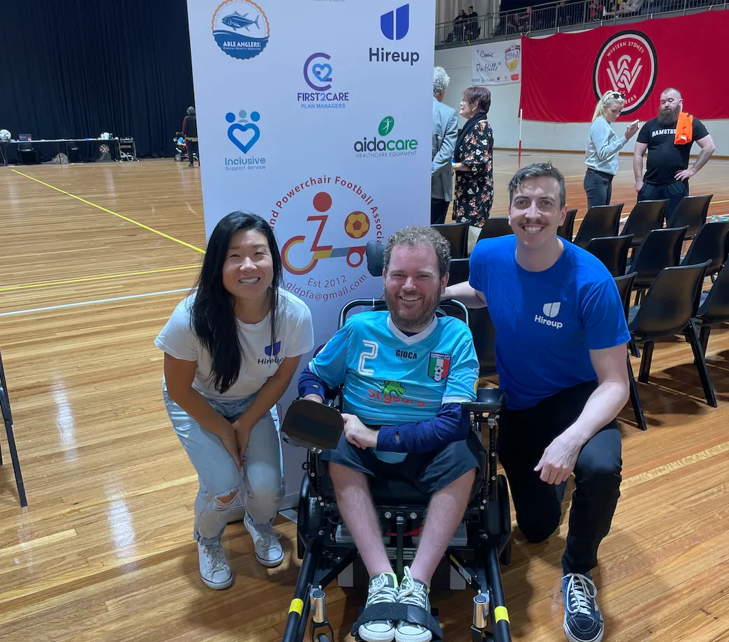 Tristram is between Steph and David from the Hireup support office. Tristram is wearing a light blue and white football jersey. Steph is wearing a white Hireup t-shirt and David is wearing a Blue Hireup t-shirt. There is a banner behind them displaying the sponsors of the Queensland powerchair football association. Tristram uses a wheelchair. Everyone is smiling.