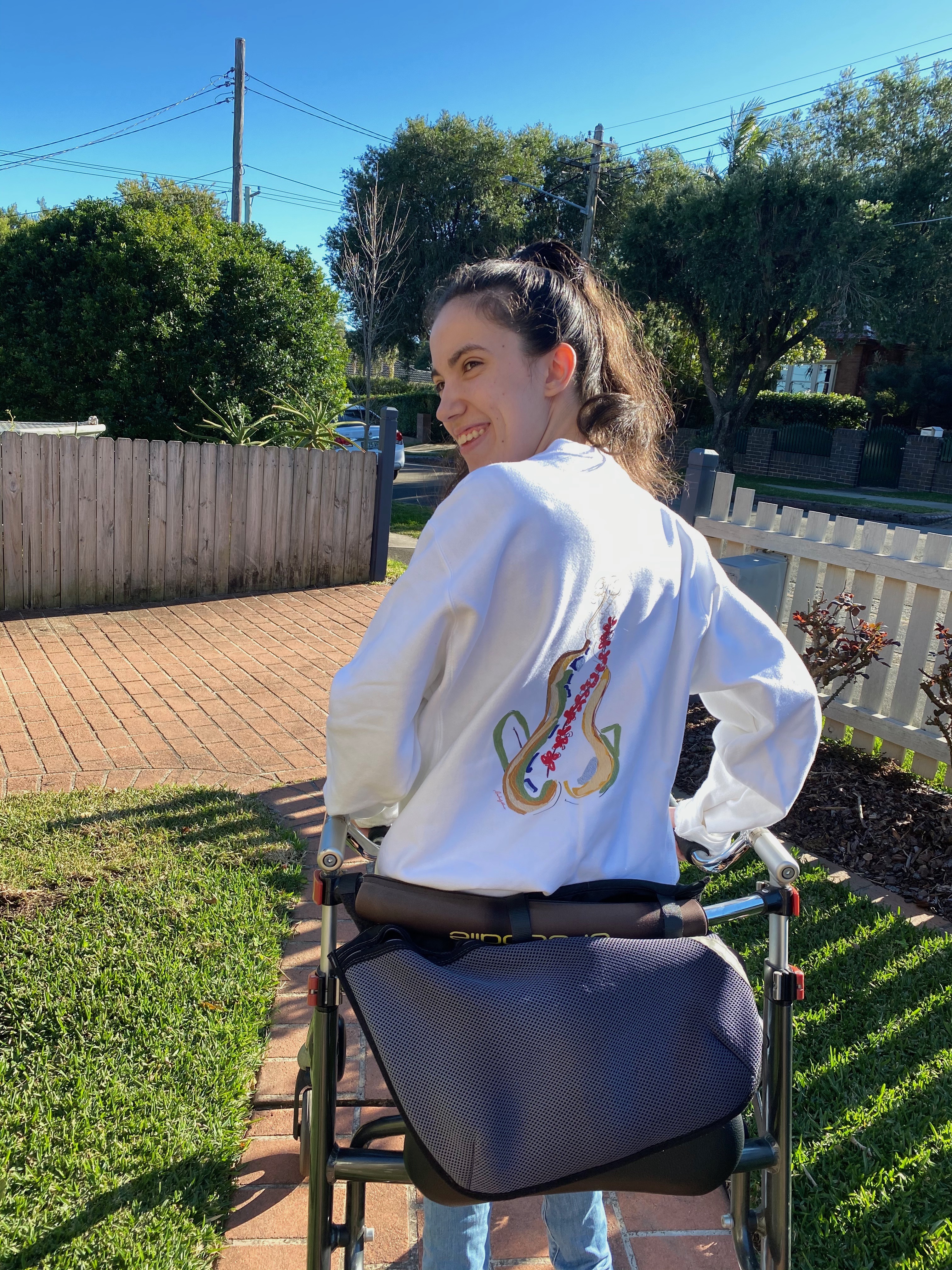 Hannah's hair is tied into a ponytail and she's wearing a jumper that has a colourful drawing depicted on it. The drawing is an outline of a woman and a branch of flowers used to represent scoliosis of the spine. Hannah is resting on her walker. 