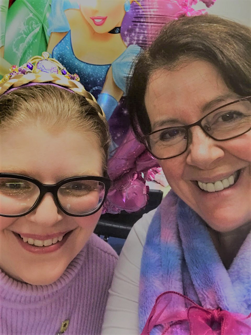Libby and Shari both smile and wear glasses. They're taking a selfie. Behind them is a colourful balloon. 