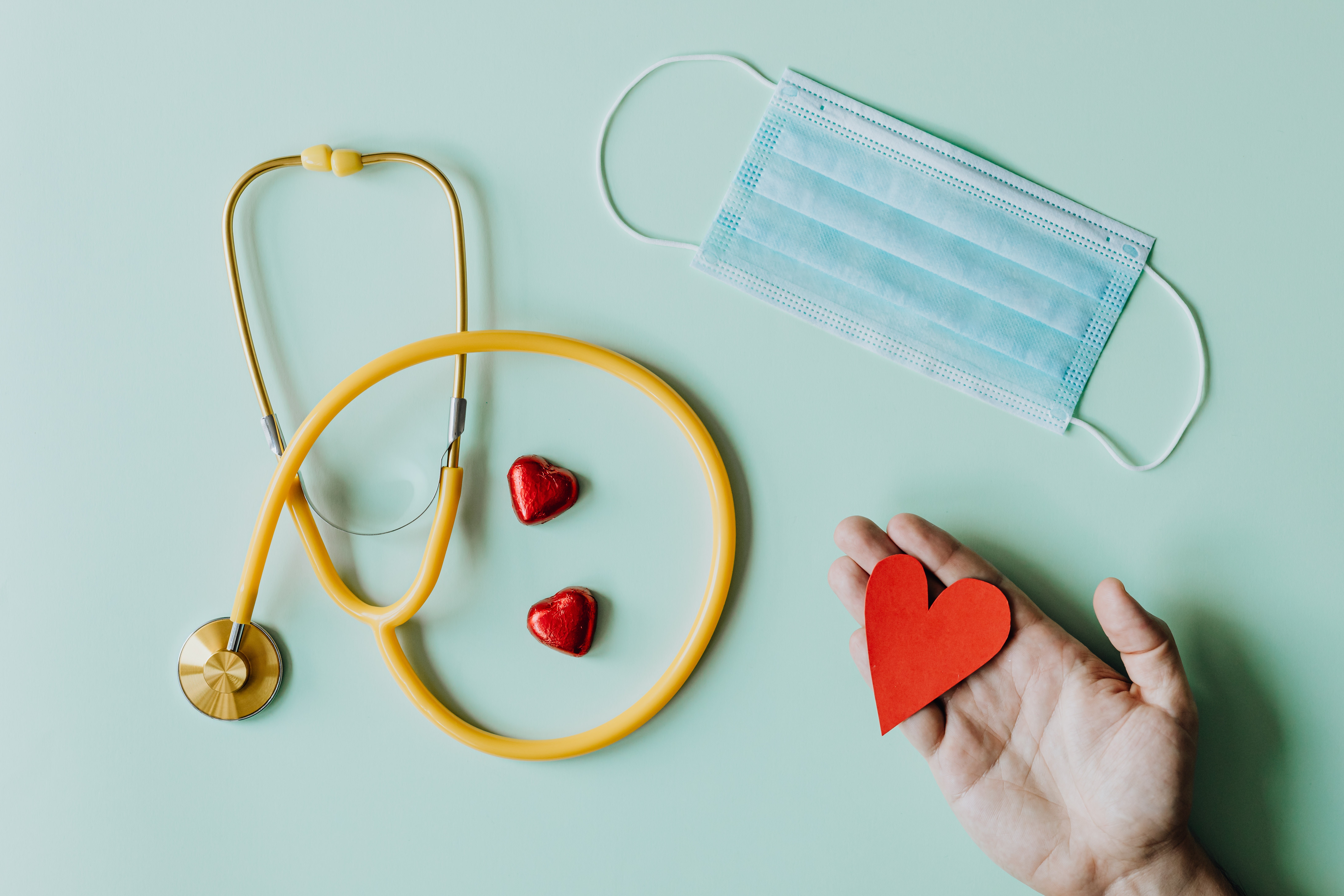 A flat lay featuring a yellow stethoscope, red chocolate hearts, blue mask and hand holding a red paper heart. 
