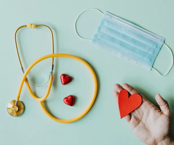 A flat lay featuring a yellow stethoscope, red chocolate hearts, blue mask and hand holding a red paper heart.