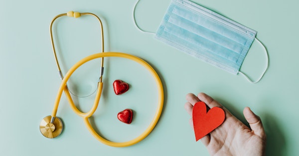 A flat lay featuring a yellow stethoscope, red chocolate hearts, blue mask and hand holding a red paper heart.