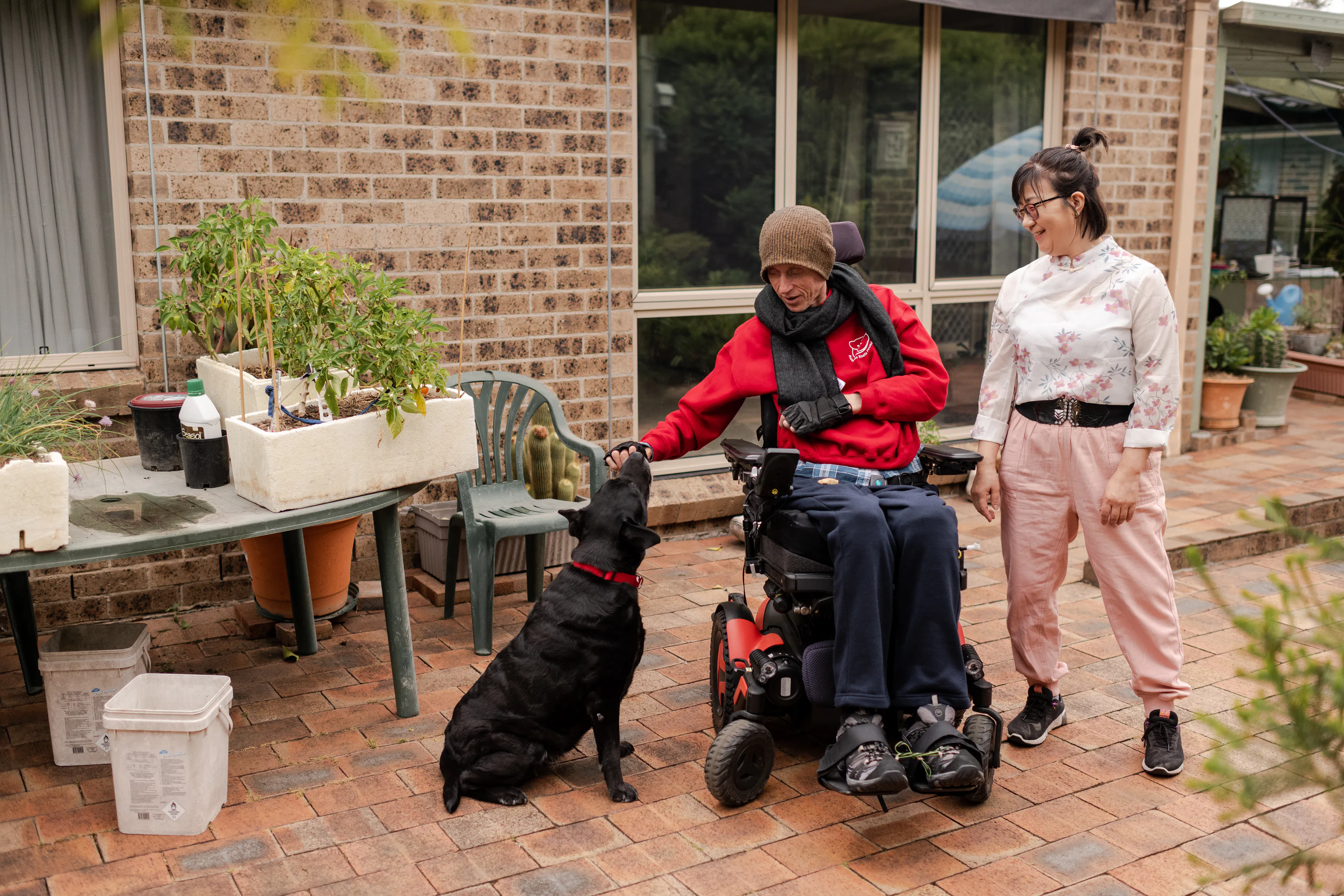Annie and Stew are smiling together on a footpath with Stew's black labrador. Annie has short black hair, wears glasses and is wearing a white top with pink flowers and pink shorts. Stew is wearing a brown beanie, black scarf, red jumper and navy track pants. Stew is a wheelchair user.