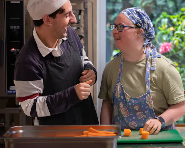 Amy and Zak are wearing aprons and chef hats. They're standing at a kitchen bench, laughing. Chopped carrots are scattered over a cutting board and are lining a baking tray.