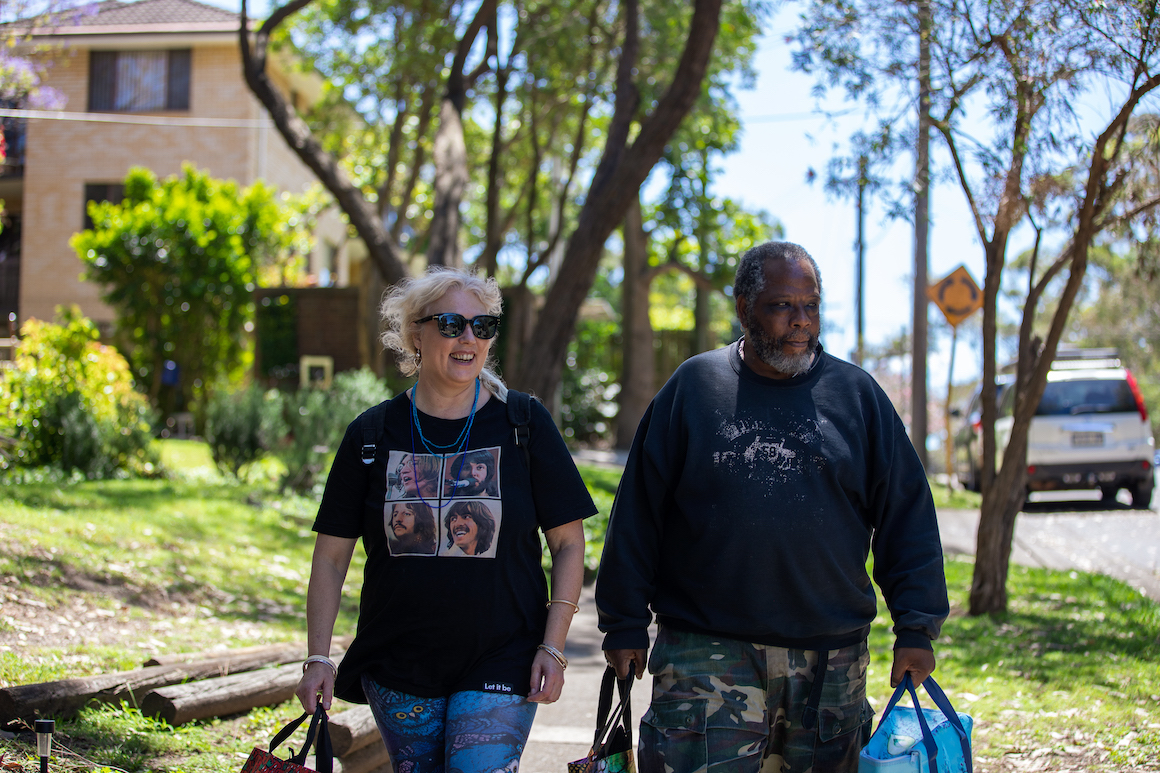 Two people are walking on a suburban footpath carrying grocery bags. The sun is shining through the trees.