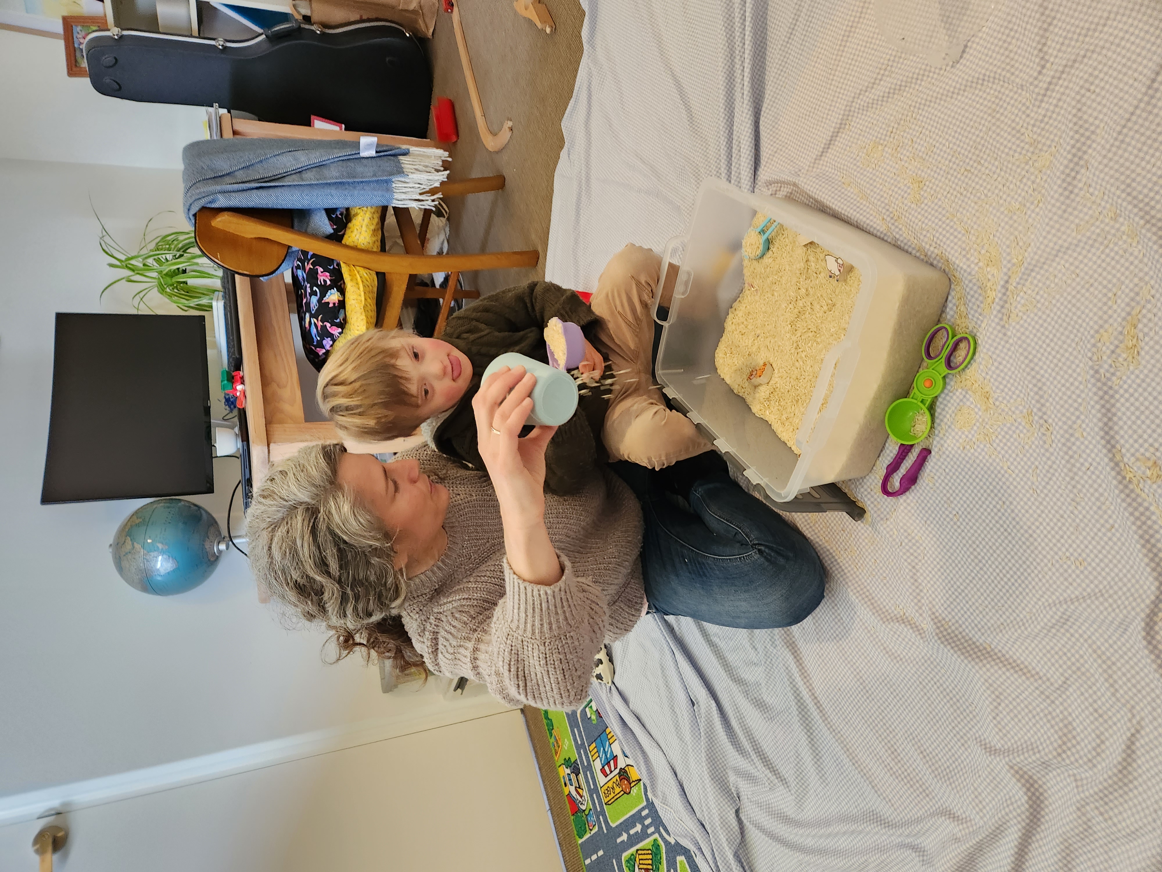 Oscar and Naomi are playing with a box of sand. Oscar has light brown hair and is wearing a brown jumper. Naomi has grey hair and is wearing a grey jumper and blue denim jeans. Naomi is pouring sand from a light blue cup.