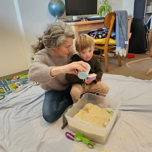 Oscar and Naomi are playing with a box of sand. Oscar has light brown hair and is wearing a brown jumper. Naomi has grey hair and is wearing a grey jumper and blue denim jeans. Naomi is pouring sand from a light blue cup.