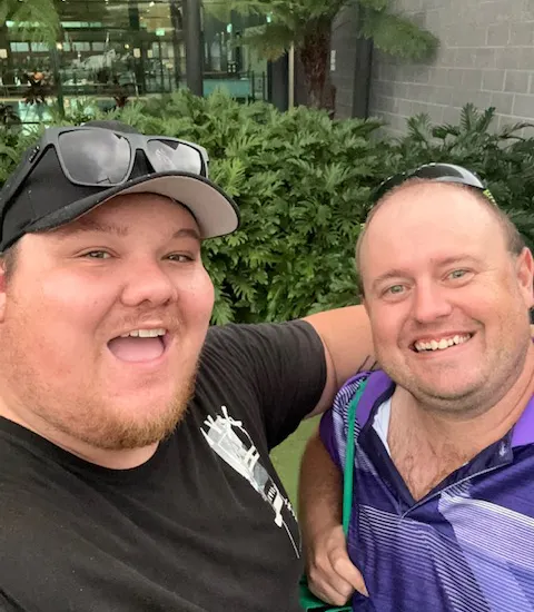 Two men wearing t-shirts and sunglasses on their head pose for a photo outside of a swimming centre.