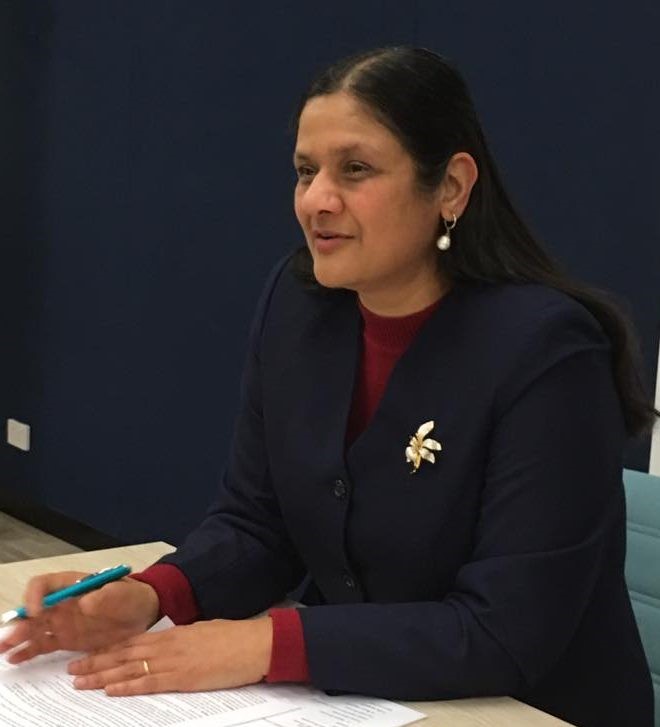Neenda Bhandari is sitting at a table. She's wearing a suit jacket and holding a pen.