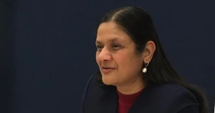 Neenda Bhandari is sitting at a table. She's wearing a suit jacket and holding a pen.