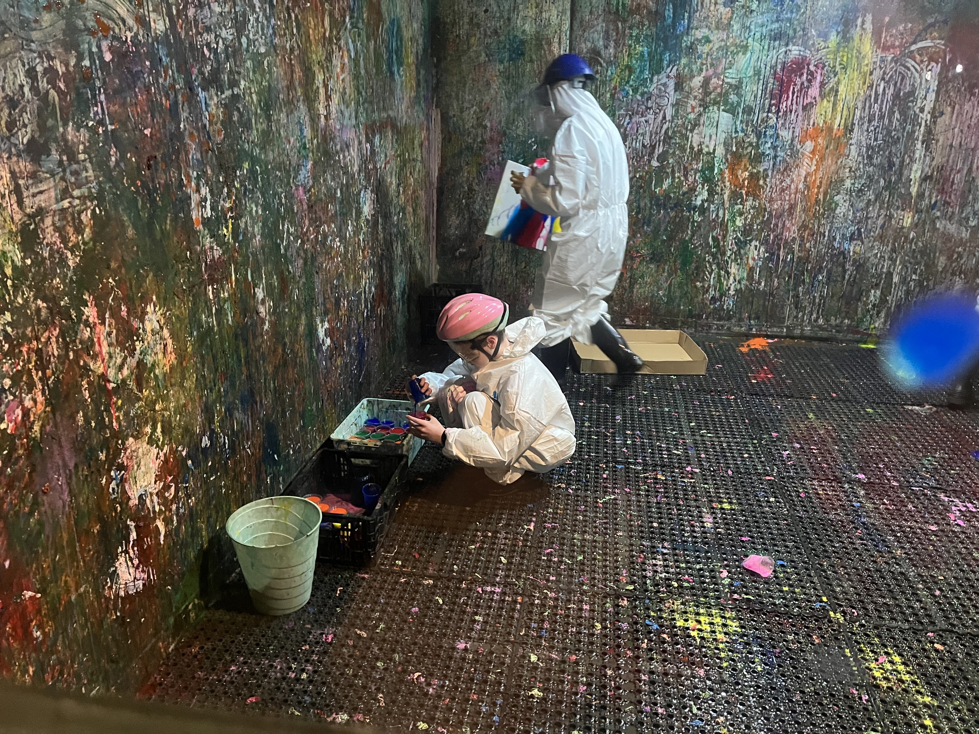 Two people wearing protective helmets and white jumpsuits are in a 'splash room' covered in paint. 