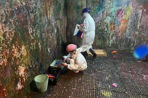 Two people wearing protective helmets and white jumpsuits are in a 'splash room' covered in paint.