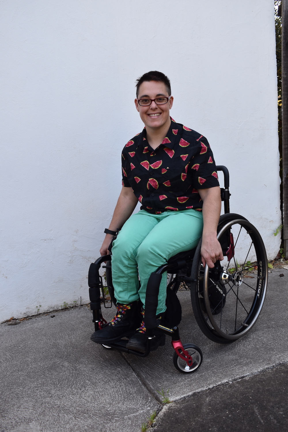 Jax is smiling toward the camera. They're wearing a shirt that has a watermelon print. Jax is a wheelchair user.  