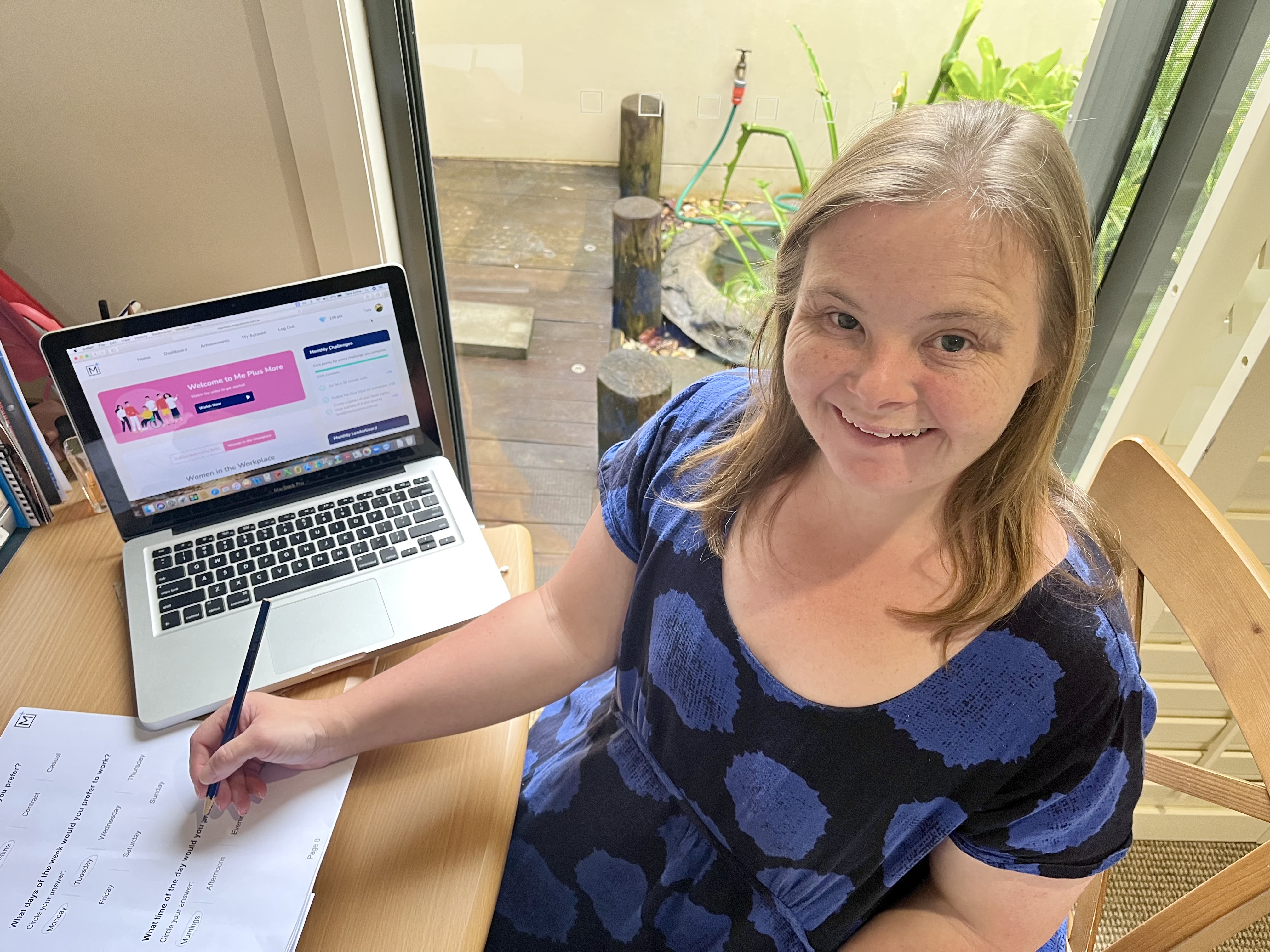 Tara is at her desk using a laptop and workbook. She is wearing a black and blue spotted dress and is smiling.