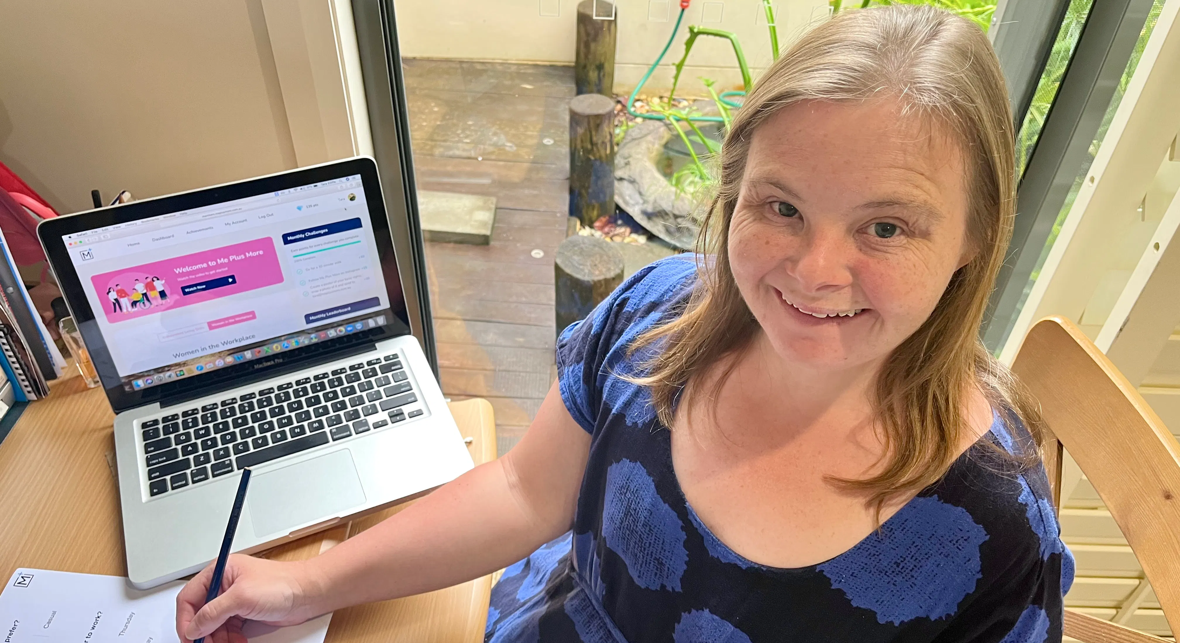 Tara is at her desk using a laptop and workbook. She is wearing a black and blue spotted dress and is smiling.