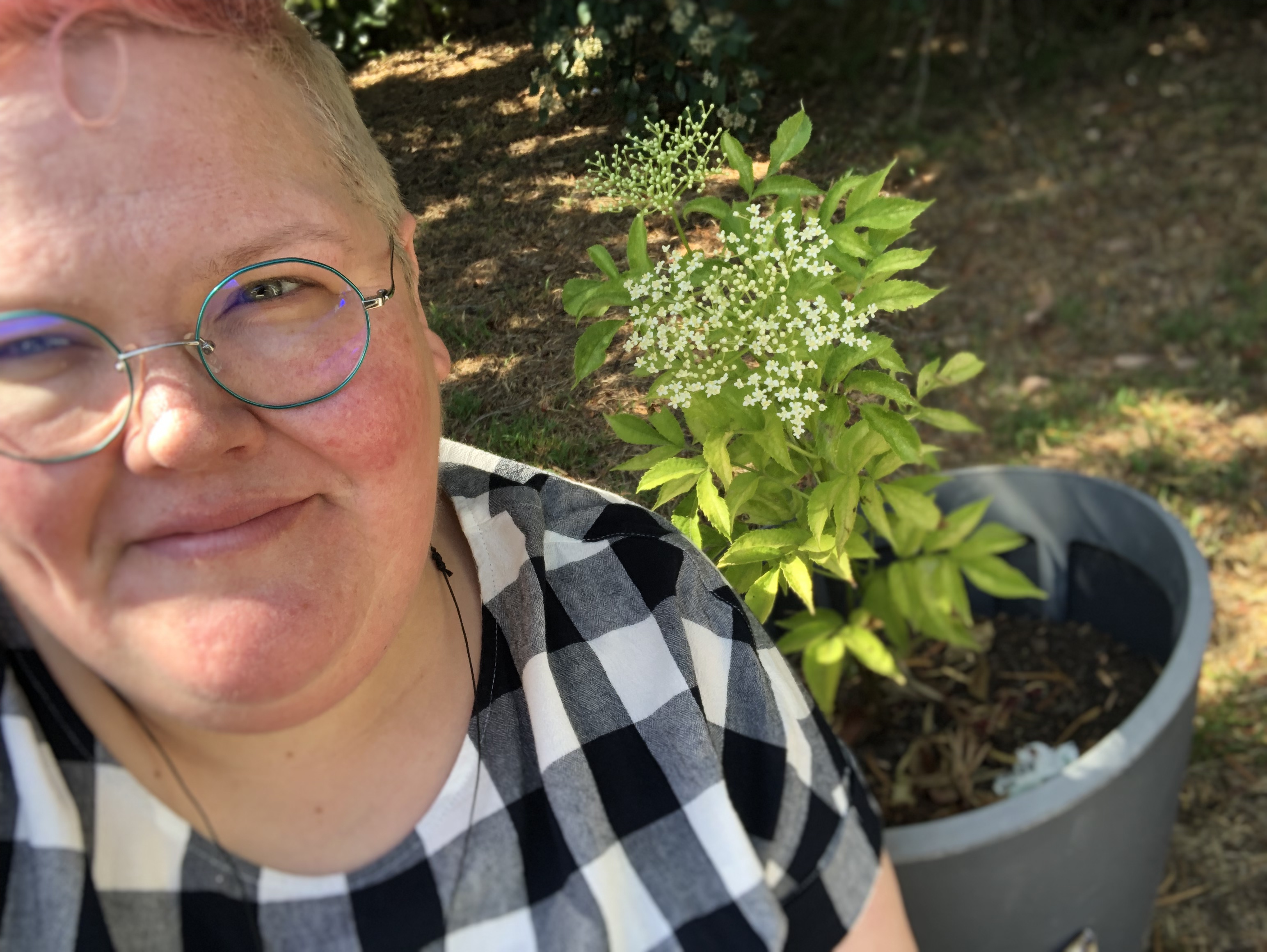 Jasper wears a shirt with check pattern and glasses. A pot plant with fine, dainty flowers is beside them. They're taking a selfie and smile gently. 