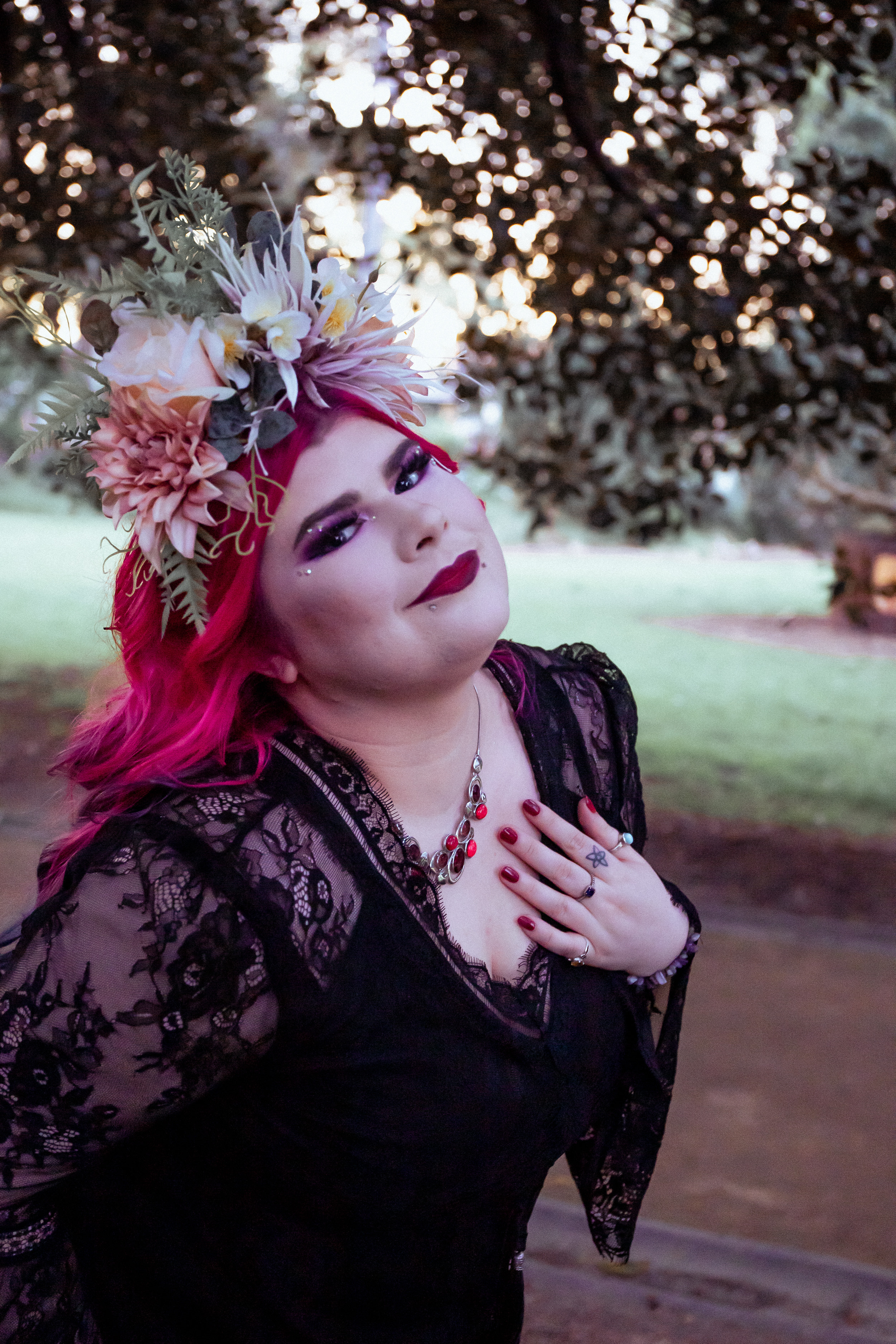 Zoe Simmons has long, bright pink hair and wears a flower crown and glamorous makeup in a park setting. She wears a black lace dress and holds her hand to her heart as she smiles at the camera. 