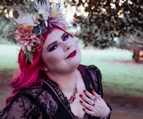 Zoe Simmons has long, bright pink hair and wears a flower crown and glamorous makeup in a park setting. She wears a black lace dress and holds her hand to her heart as she smiles at the camera.
