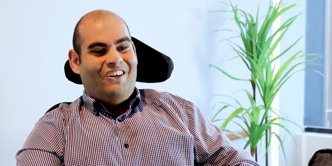 Elie wears a check shirt and smiles. He's in an office. Elie is a wheelchair user. 