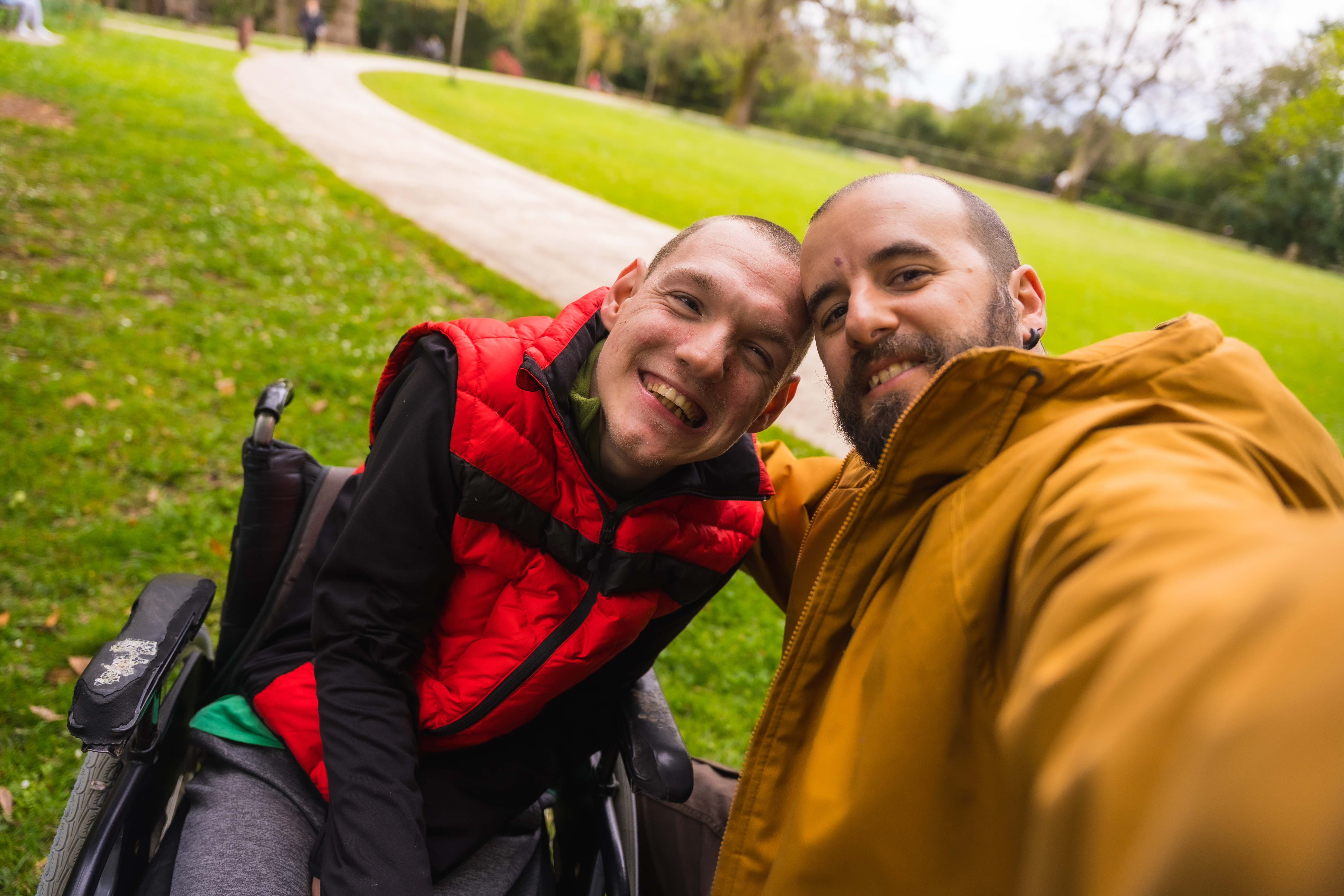 Two men with shaved short brown hair are smiling. One is wearing a bright red puffer vest, black long sleeved shirt, grey tracksuit pants and is a wheelchair user. The other man is wearing a mustard yellow rain jacket. There is a park with short green grass and a footpath in the background.