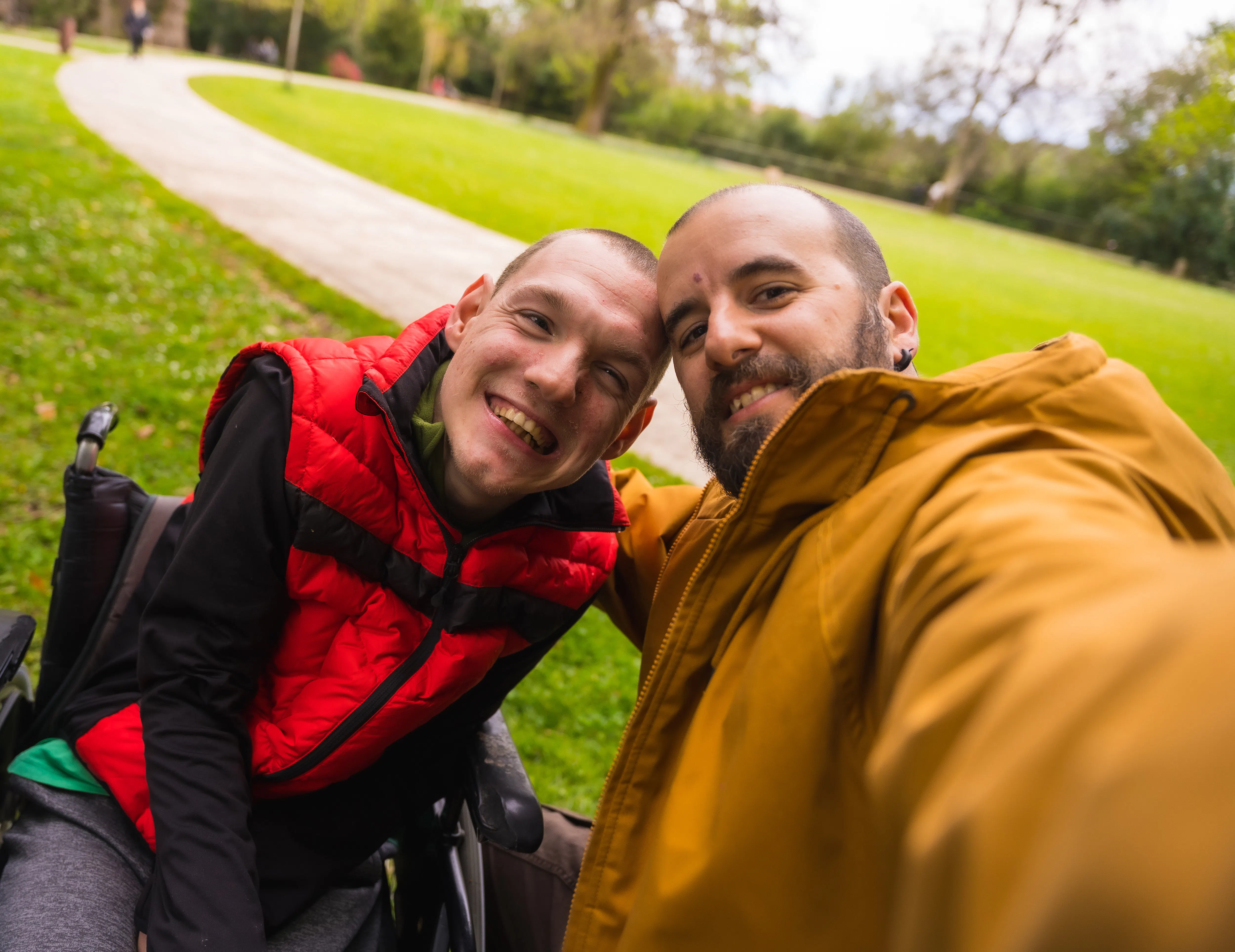 Two men with shaved short brown hair are smiling. One is wearing a bright red puffer vest, black long sleeved shirt, grey tracksuit pants and is a wheelchair user. The other man is wearing a mustard yellow rain jacket. There is a park with short green grass and a footpath in the background.