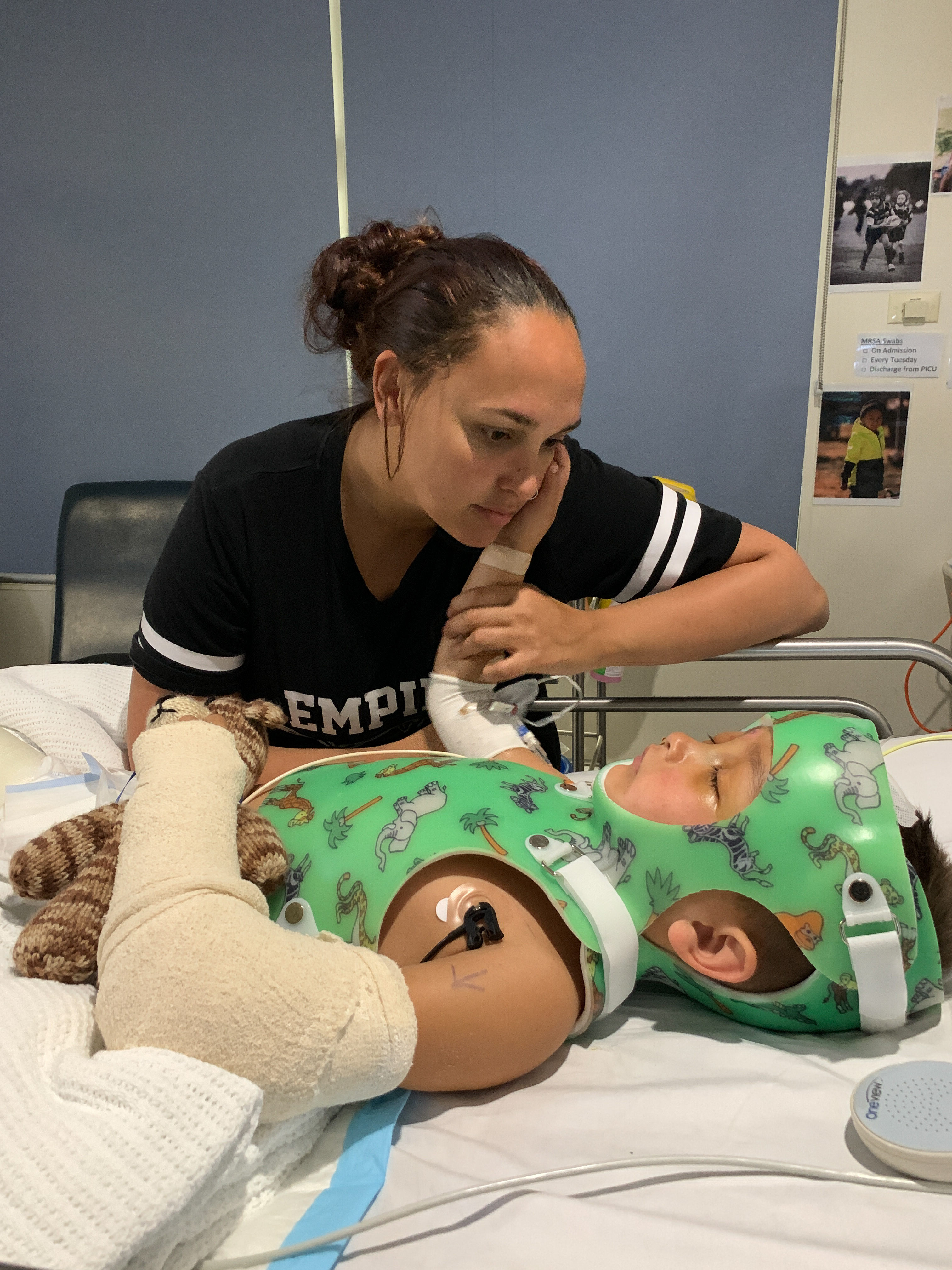 Durakai is in a body cast, he has monitors attached to his body and his arms are wrapped in dressings. His mum, Brinae, is watching over him from the side of the bed. Durakai has his right arm raised, touching his mum on the cheek. 
