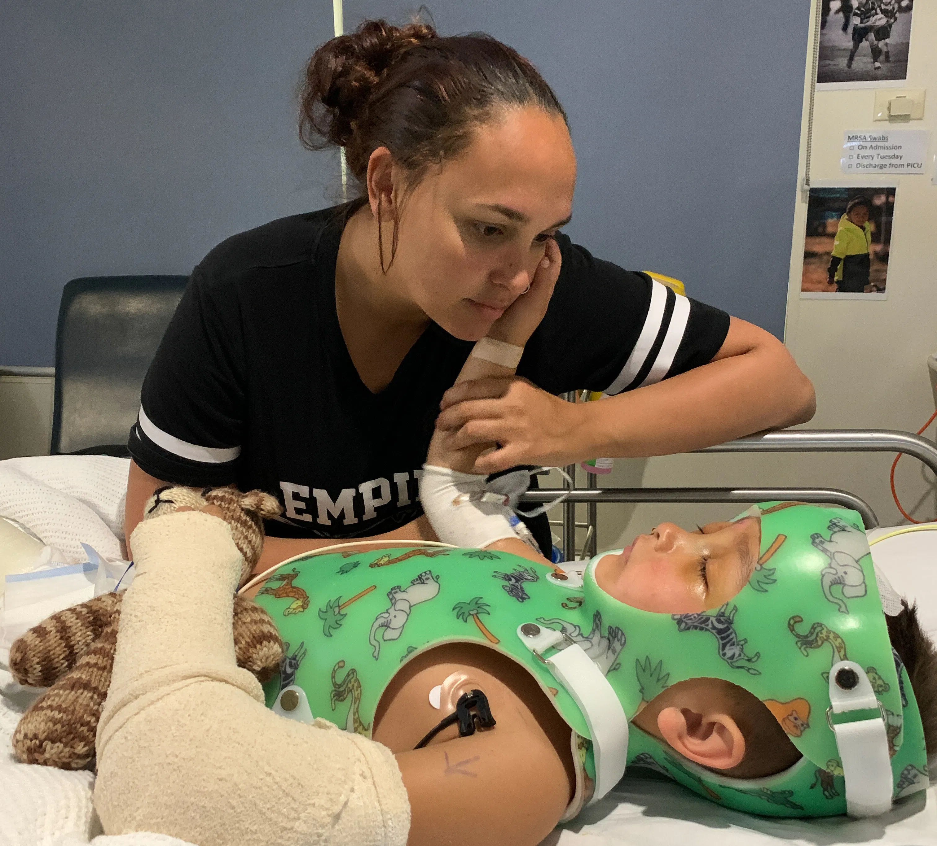Durakai is in a body cast, he has monitors attached to his body and his arms are wrapped in dressings. His mum, Brinae, is watching over him from the side of the bed. Durakai has his right arm raised, touching his mum on the cheek.