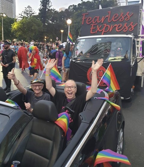 Image shows Dr David Abello in the back of a black convertible at the Mardi Gras parade in 2019. The car is decked with rainbow flags. David is beaming at the camera wit his hands in the air. Photo is courtesy of Sallie Colechin.