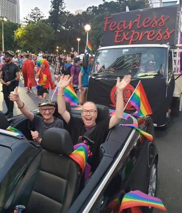 Image shows Dr David Abello in the back of a black convertible at the Mardi Gras parade in 2019. The car is decked with rainbow flags. David is beaming at the camera wit his hands in the air. Photo is courtesy of Sallie Colechin.