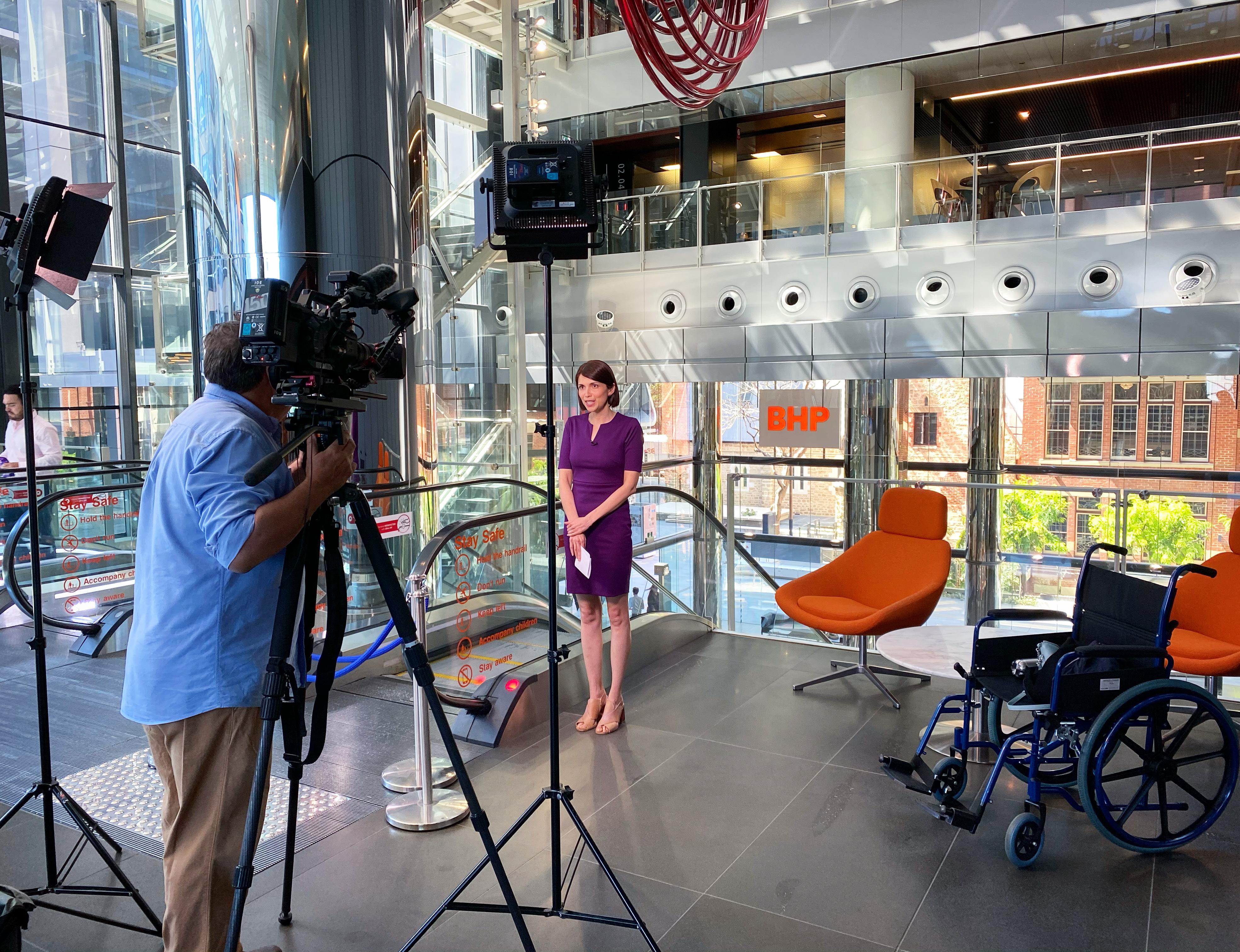 Natalia Hodgins stands in a news studio and talks to a camera, held by a cameraman. There are two chairs and a wheelchair to the side of the photo. 