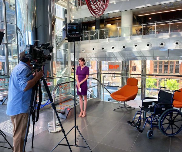 Natalia Hodgins stands in a news studio and talks to a camera, held by a cameraman. There are two chairs and a wheelchair to the side of the photo.