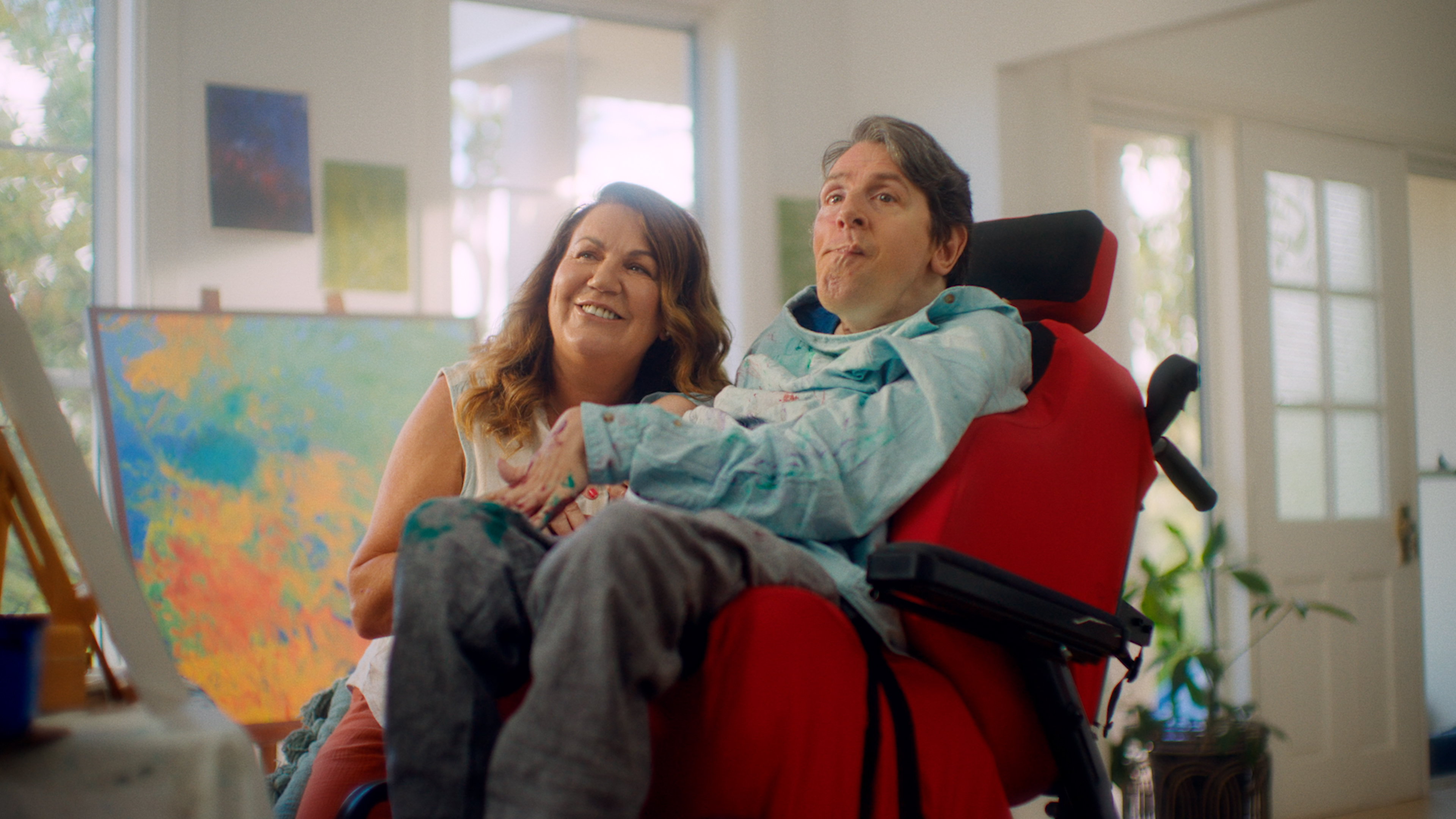 Glenn and Fadwa are looking at an art canvas. Fadwa has light brown hair and is smiling. Glenn uses a bright red wheelchair and is wearing a light blue painting smock.