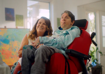 Glenn and Fadwa are looking at an art canvas. Fadwa has light brown hair and is smiling. Glenn uses a bright red wheelchair and is wearing a light blue painting smock.