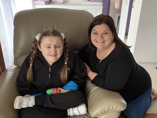 Jessica is sitting cross-legged on an armchair. Kneeling beside her is Donna, her support worker. They're both smiling. 