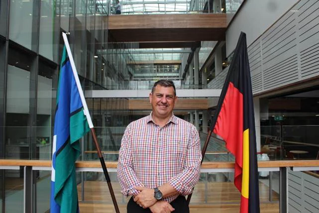 Scott Avery is wearing a red and white chequered shirt and is smiling. He is standing in front of the Torres strait islander flag and the Indigenous Australian flag.
