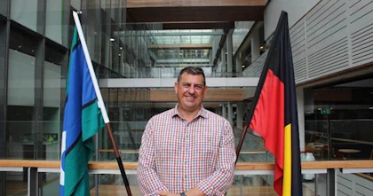 Scott Avery is wearing a red and white chequered shirt and is smiling. He is standing in front of the Torres strait islander flag and the Indigenous Australian flag.