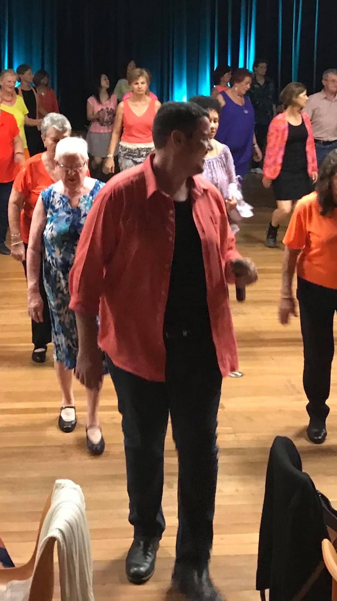 Josh is dancing at the front of the line dancing class. He is wearing a salmon coloured shirt and black pants.