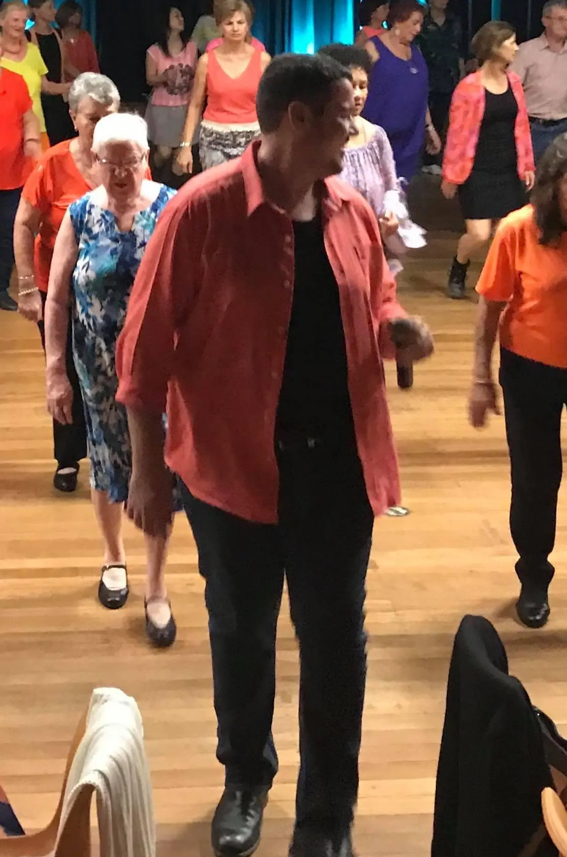 Josh is dancing at the front of the line dancing class. He is wearing a salmon coloured shirt and black pants.