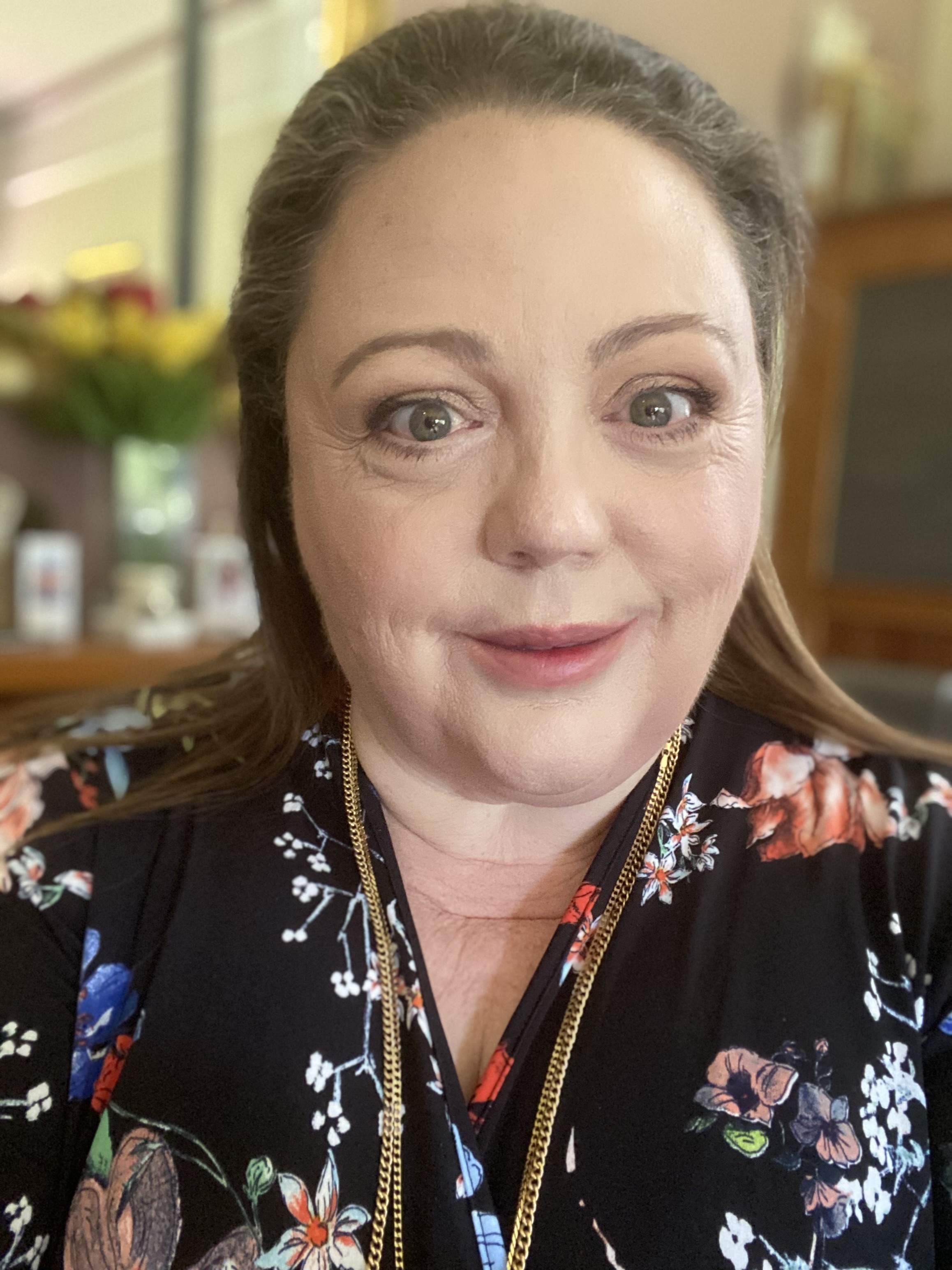 Kate Lindsay has long brown hair, green eyes and fair skin. She wears a black, floral-printed top and gold necklaces. 
