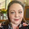 Kate Lindsay has long brown hair, green eyes and fair skin. She wears a black, floral-printed top and gold necklaces.