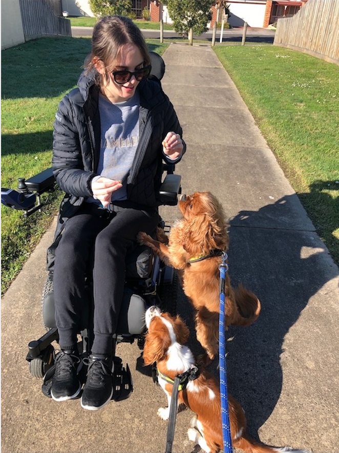 Tayla is playing with two dogs. She has brown hair and is wearing sunglasses. Tayla is wearing black runners, black track-pants, a grey top and a black puffer jacket. Tayla uses a wheelchair. Two are jumping up on the side of her wheelchair.