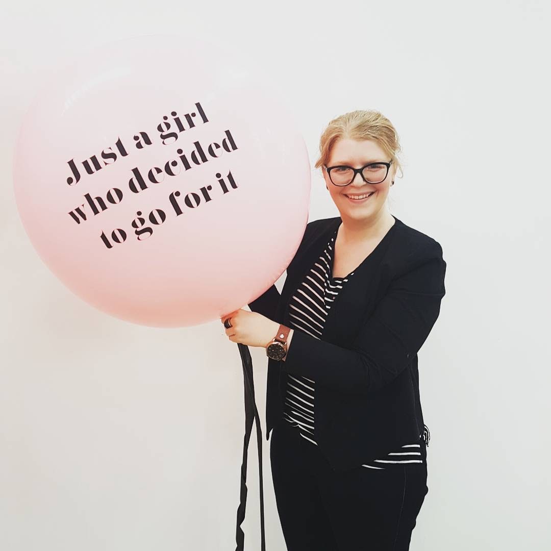 Libby is smiling and is holding onto a large pink balloon that has the words 'Just a girl who decided to go for it' printed on the balloon. Libby wears glasses and a dark coloured cardigan. 