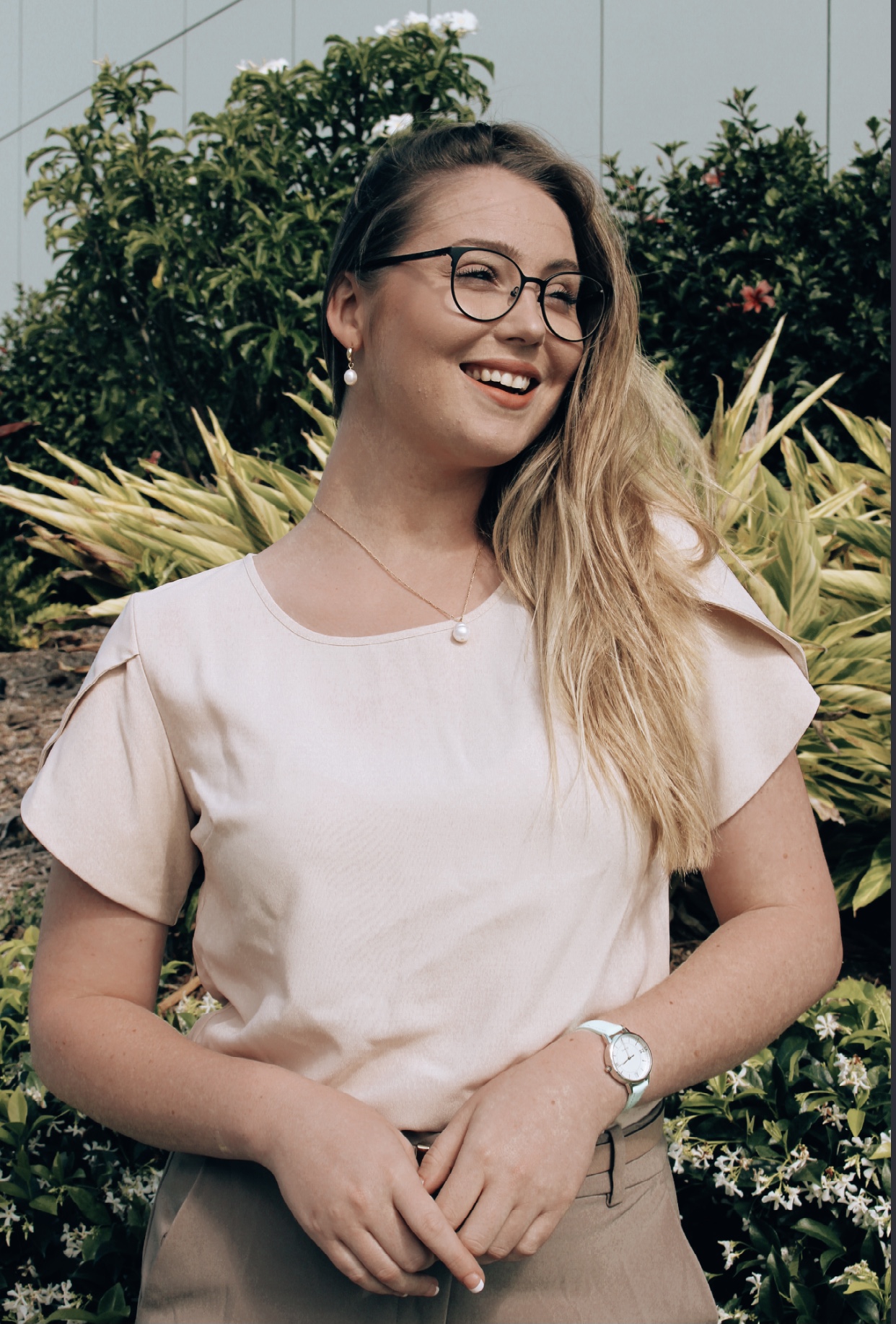 Megan looks into the distance and a garden is behind her. Megan has wears glasses and she wears a light coloured top and trousers, and a pearl necklace. 