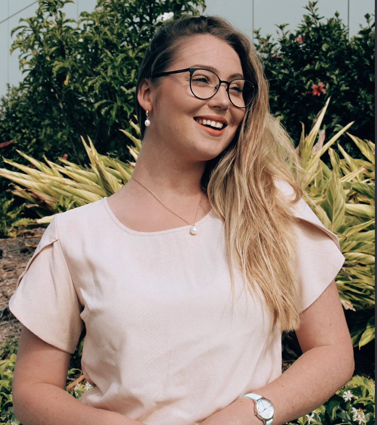 Megan looks into the distance and a garden is behind her. Megan has wears glasses and she wears a light coloured top and trousers, and a pearl necklace.