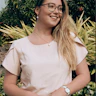 Megan looks into the distance and a garden is behind her. Megan has wears glasses and she wears a light coloured top and trousers, and a pearl necklace.